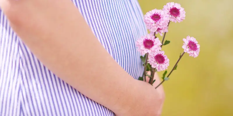 O femeie tanara insarcinata se tine cu mana de burtica, cu flori roz-rosii in mana