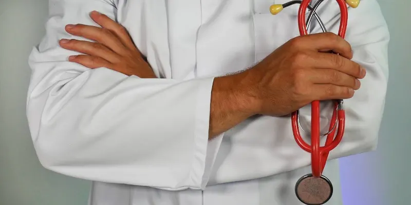 Doua maini ale unui medic in halat alb, incrucisate la piept, cu un stetoscop rosu in mana stanga
